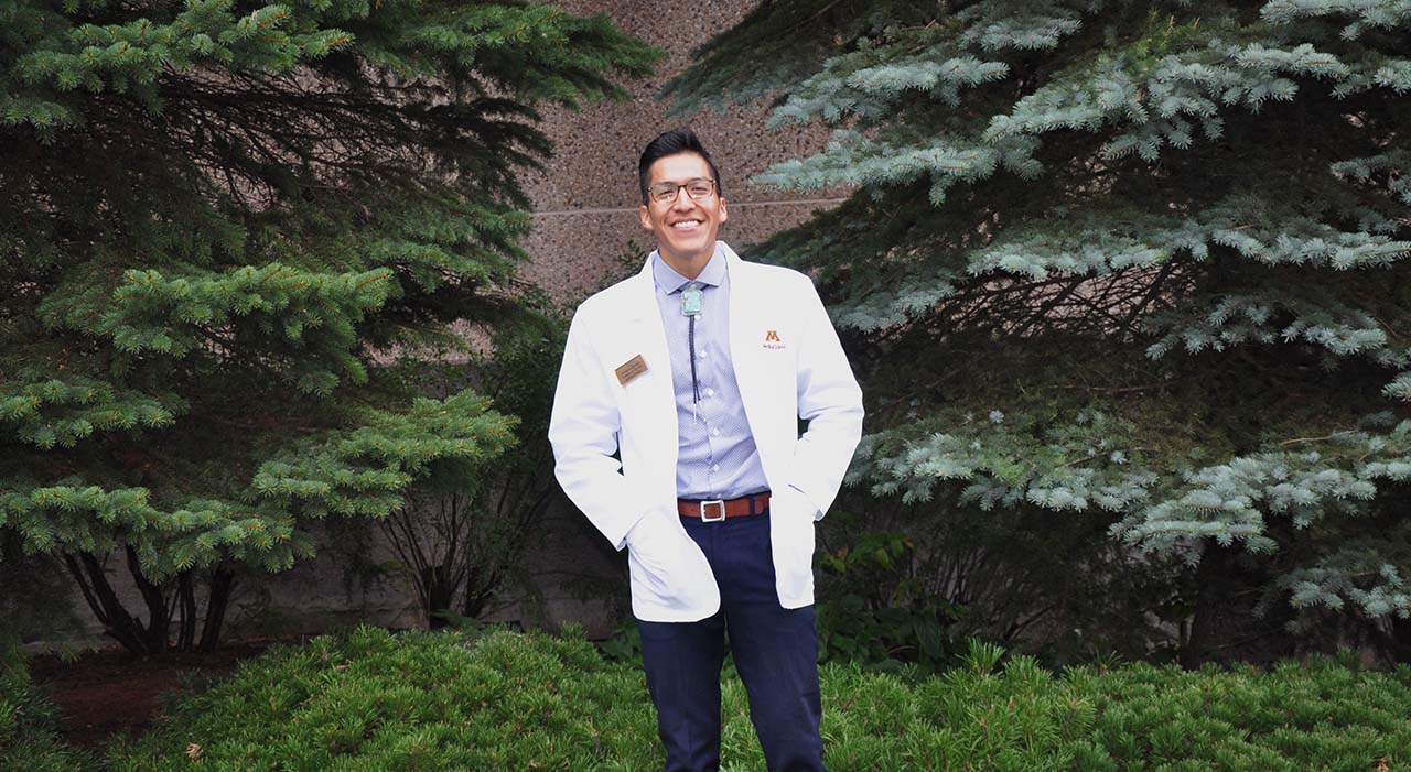 Photo of Casey Smith, med student, posing for a photo in front of some evergreen trees.