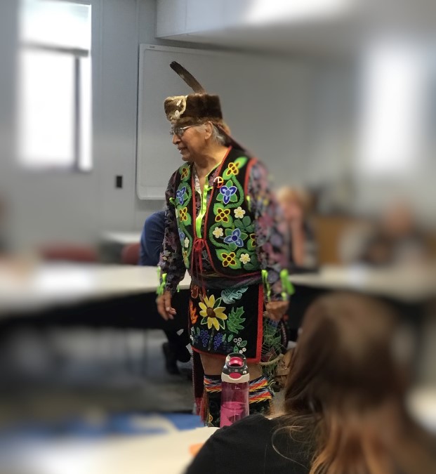 Native elder Dennis White addresses room dressed in regalia.
