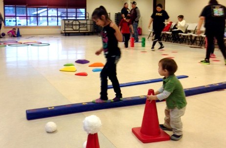 A toddler carries a red cone while observing an older child walking on the balance beam
